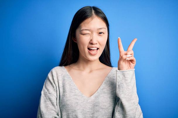 Jeune belle femme asiatique portant un pull décontracté debout sur fond bleu isolé souriant avec un visage heureux clin d'oeil à la caméra faisant signe de victoire avec les doigts. Numéro deux
. - Photo, image