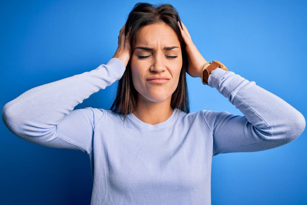 Jeune belle femme brune portant un pull décontracté debout sur fond bleu souffrant de maux de tête désespérés et stressés parce que la douleur et la migraine. Mains sur la tête
. - Photo, image