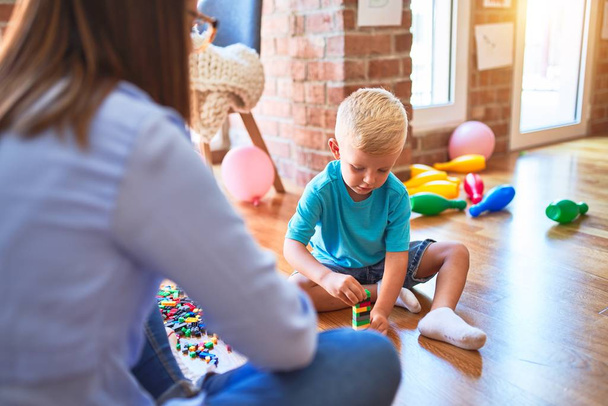 Young caucasian child playing at playschool with teacher. Mother and son at playroom bulding a tower with toy blocks - Photo, Image