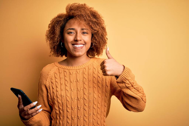 Young African American afro woman with curly hair having conversation using smartphone happy with big smile doing ok sign, thumb up with fingers, excellent sign - 写真・画像