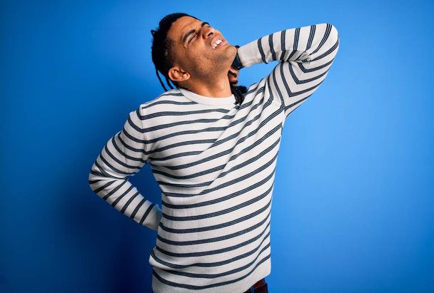 Junger gut aussehender afro-amerikanischer Afro-Mann mit Dreadlocks, der lässig gestreifte Pullover trägt Leiden an Nackenschmerzen, Berührung des Nackens mit der Hand, Muskelschmerzen - Foto, Bild