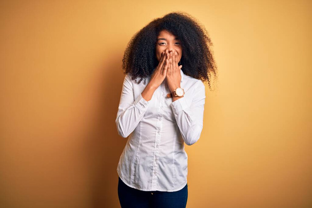 Young beautiful african american elegant woman with afro hair standing over yellow background laughing and embarrassed giggle covering mouth with hands, gossip and scandal concept - Foto, immagini