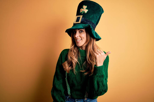 Beautiful brunette woman wearing green hat with clover celebrating saint patricks day smiling with happy face looking and pointing to the side with thumb up. - Foto, Imagem