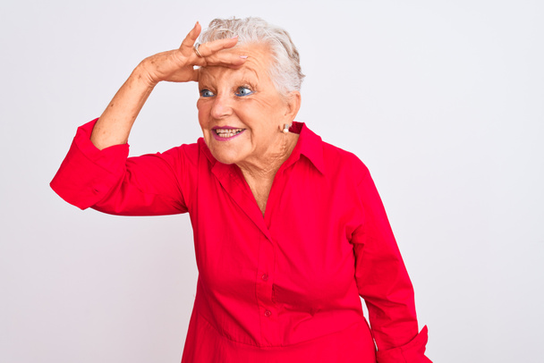 Eine ältere grauhaarige Frau in rotem lässigem Hemd steht vor isoliertem weißem Hintergrund sehr glücklich und lächelt mit der Hand über dem Kopf in die Ferne. Suchkonzept. - Foto, Bild