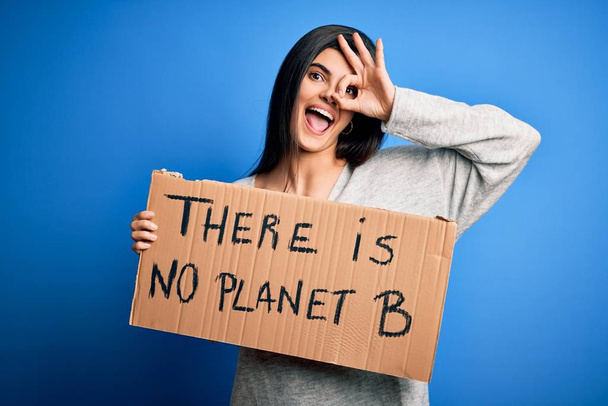 Jeune belle militante brune tenant une bannière protestant pour prendre soin de la planète avec un visage heureux souriant faisant ok signe avec la main sur les yeux regardant à travers les doigts
 - Photo, image