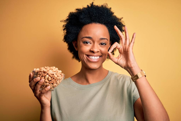 Fiatal afro afro nő göndör haj gazdaság tál sós mogyoró csinál rendben jel ujjakkal, kiváló szimbólum - Fotó, kép