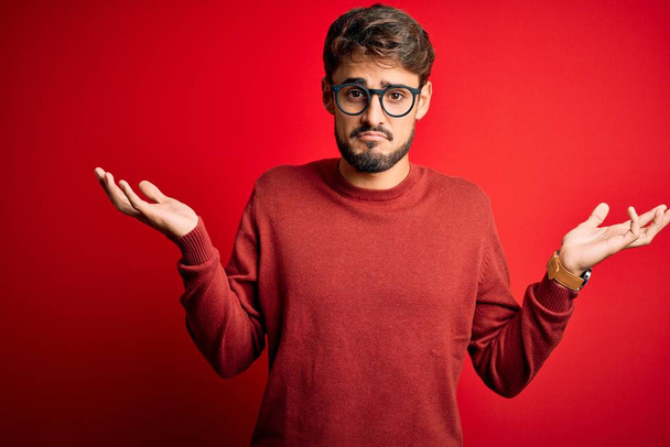 Fiatal jóképű férfi szakállal szemüvegben és pulóverben áll a piros háttér felett tudatlan és zavaros kifejezés karokkal és felemelt kézzel. Kétséges fogalom. - Fotó, kép