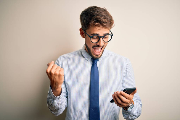 Young businessman having a conversation using smartphone over white background screaming proud and celebrating victory and success very excited, cheering emotion - 写真・画像