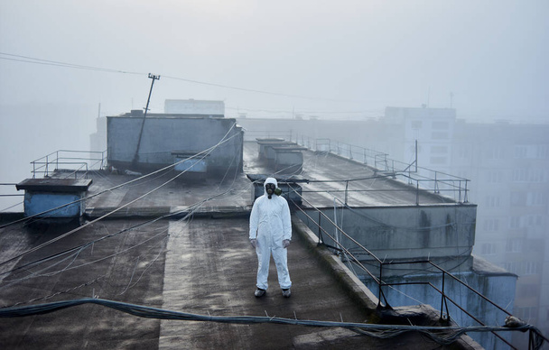 Hombre científico de pie en el techo de un edificio de planta alta, con una bata blanca y máscara de gas, explorando el territorio para futuras investigaciones
 - Foto, Imagen