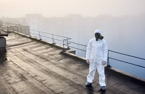 Man wearing white coverall and gas mask, standing on the roof of an urban building with his back to a rising sun in foggy weather, concept of environmental problem - Fotografie, Obrázek