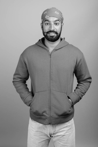 Студийный снимок молодого красивого бородатого индийца на сером фоне в черно-белом - Фото, изображение