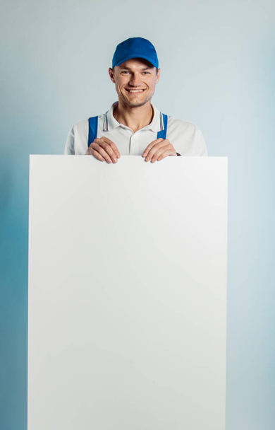 Imagen burlona de un joven trabajador oloroso sosteniendo una pancarta blanca vacía. Fondo blanco o azul. Concepto empresarial
. - Foto, imagen
