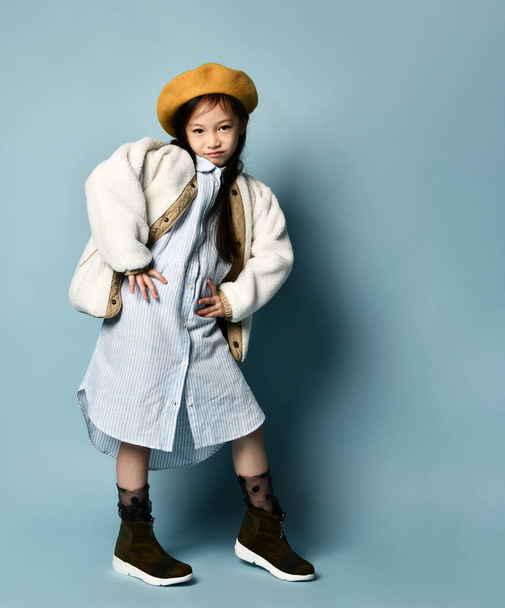 Little asian schoolgirl in shirt dress, double sided jacket, brown beret, boots. Smiling, posing on blue background. Full length - Photo, Image