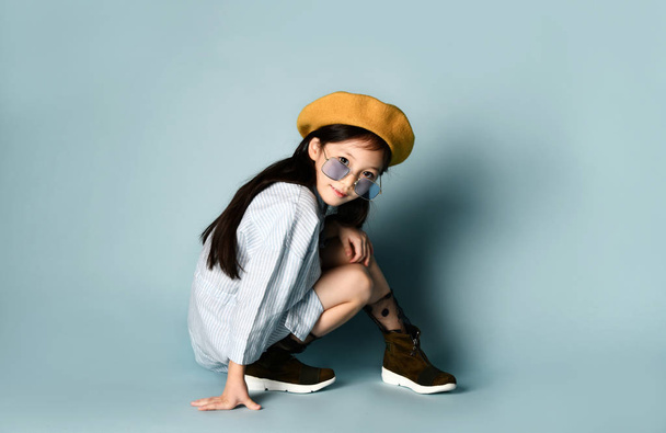 Little asian child in sunglasses, shirt dress, brown beret, boots. Smiling, posing sitting on blue background. Close up - Photo, image