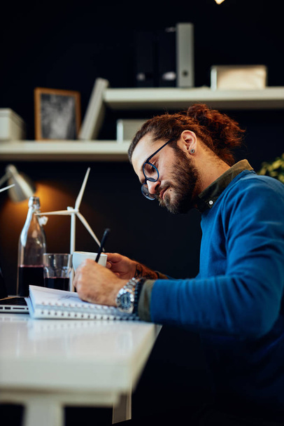 Νέος καινοτόμος, αφοσιωμένος γενειοφόρος εργαζόμενος κάθεται στο σύγχρονο γραφείο του και σχεδιάζει σκίτσα ανεμόμυλων και πίνοντας καφέ. Έννοια της αειφόρου ανάπτυξης. - Φωτογραφία, εικόνα