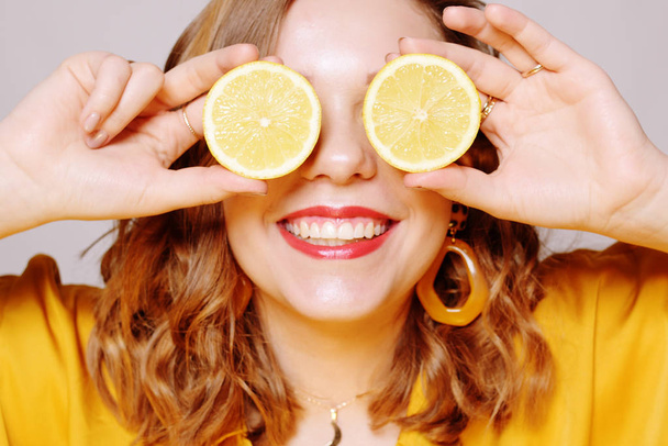 Clouse up of a girl with a lemon. A girl in a bright yellow dress, with yellow earrings, holds a lemon. Sincere smile. covering eye with limon. - Fotografie, Obrázek