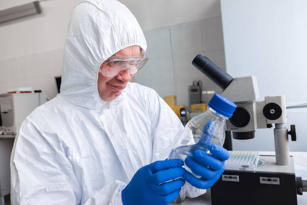 Een bioloog in een wit beschermend pak en bril werkt met een microscoop in het laboratorium. Het concept van epidemie en pandemie - Foto, afbeelding
