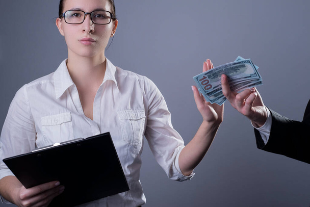 Portret van een zakenmeisje in een bril met serieuze emoties op haar gezicht, met een hand die verbiedt haar dollarbiljetten van een man te geven, tegen de achtergrond van steekpenningen. Studio foto op een grijze achtergrond - Foto, afbeelding