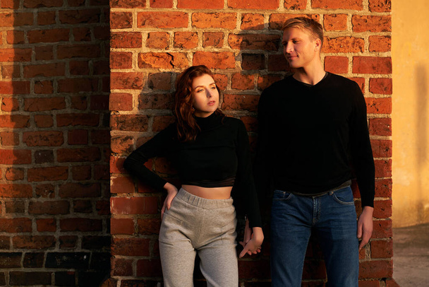Romantic couple young beautiful sensual redhead woman and tall handsome blonde man posing in sunset sunlight on red brick wall background - Φωτογραφία, εικόνα