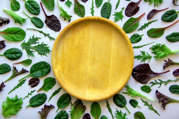 Wooden round plate, baby leaves of vegetables laid out around. Assortment with arugula, mongold, spinach, lettuce on a white background. Healthy nutrition concept. Top view, flat lay, copy space. - Photo, Image