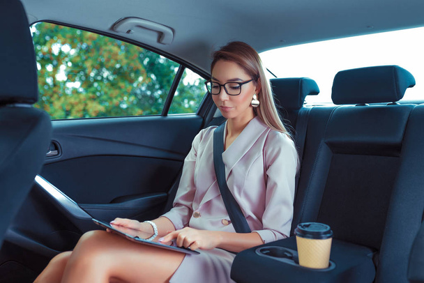 Beautiful secretary girl VIP taxi business class, works on tablet, holds touch screen, tanned figure with long hair, cup coffee tea. Work on road, business meeting in car. Summer city traffic jam. - Fotoğraf, Görsel
