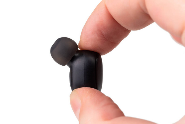 two caucasian fingers holding a black wireless earbud - close-up with selective focus on white background - Photo, Image