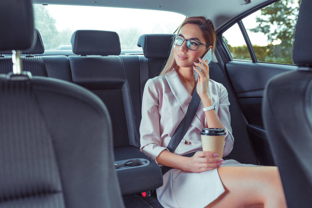 Beautiful girl secretary in business class of VIP taxi calling on phone, holding smartphone, fashionable and modern woman, cup of coffee with tea. Work on road, business meeting car. Meets passenger. - Zdjęcie, obraz