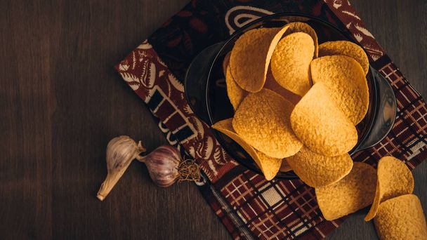 Хрустящие картофельные чипсы в миске и чеснок на деревянном фоне, с кухонным полотенцем
 - Фото, изображение