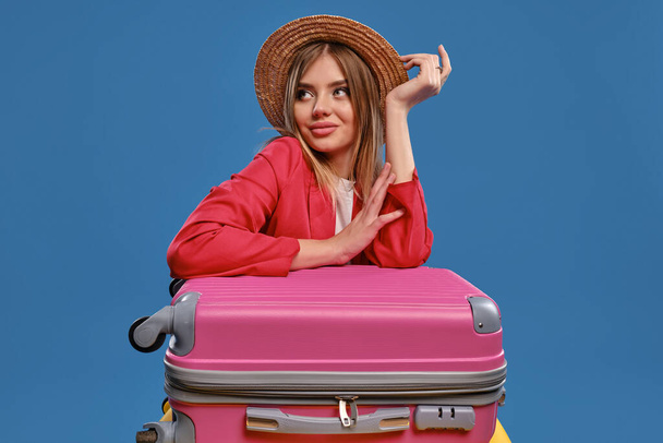 Blonde girl in straw hat, white blouse, red jacket. Sitting leaning on colorful suitcases, posing on blue background. Travelling concept. Close-up - Fotó, kép
