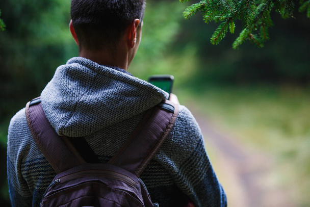 A young man with a backpack in the woods looking at the phone-a view from the back. Orientation on a walk along a forest path using the Navigator in your phone - 写真・画像
