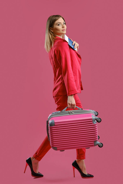Mujer rubia en traje pantalón rojo, tacones negros altos. Sonriendo, llevando maleta, con pasaporte y billete, fondo rosa. Concepto de viaje
 - Foto, imagen