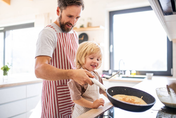 A side view of small boy with father indoors in kitchen making pancakes. - Valokuva, kuva