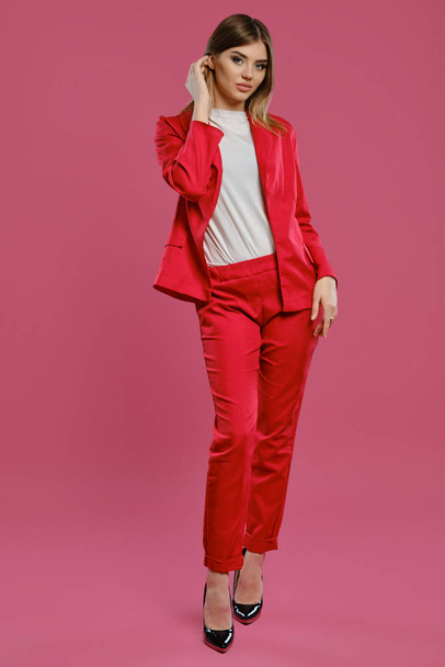 Blonde female in red pantsuit, white blouse, high black heels. Posing standing against pink studio background. Beauty, fashion. Full length - 写真・画像