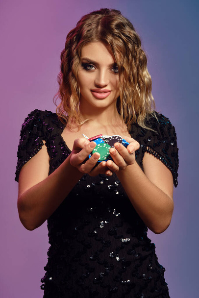 Blonde woman in black sequin dress is smiling, showing handful of multicolored chips, posing on colorful studio background. Poker, casino. Close-up. - Foto, Bild