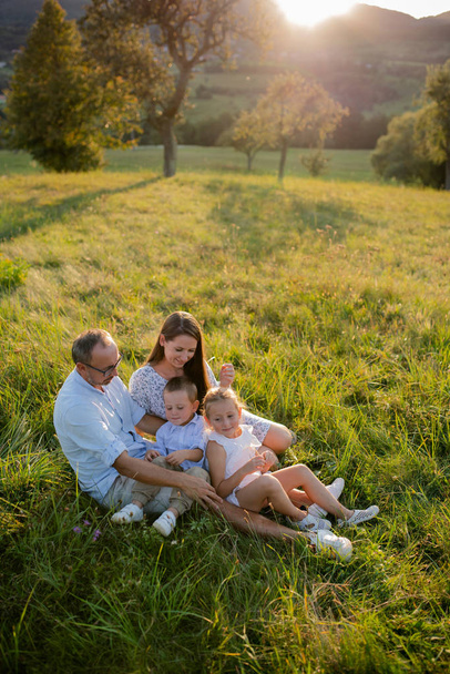 Mladá rodina se dvěma malými dětmi sedící na louce při západu slunce. - Fotografie, Obrázek