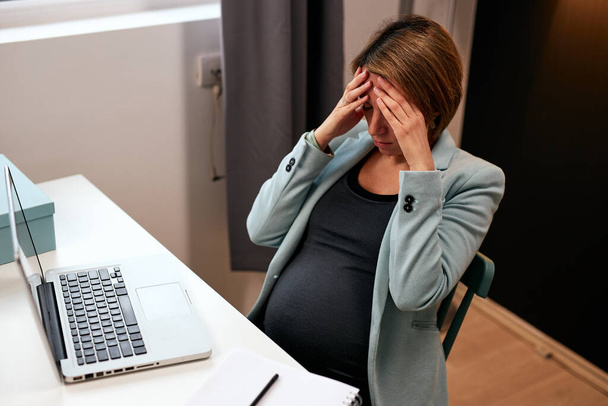 Těhotná podnikatelka ve čtyřicátých letech sedí ve své kanceláři a bolí ji hlava. - Fotografie, Obrázek