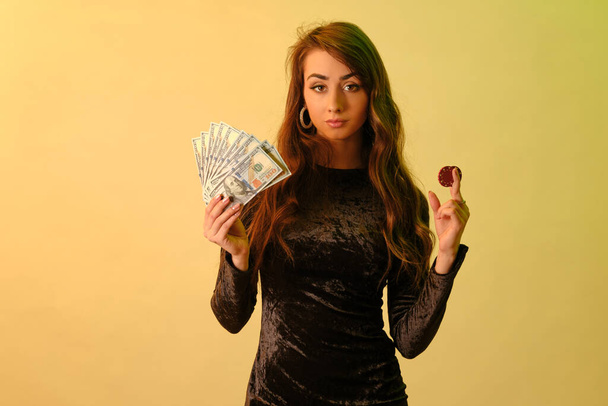 2つの赤いチップといくつかのお金を示す黒のドレスのブルネットの女の子は、黄色のスタジオの背景に対してポーズ。ギャンブル、ポーカー、カジノ。閉鎖. - 写真・画像