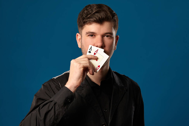 Newbie in poker, in black vest and shirt. Holding two playing cards while posing against blue studio background. Gambling, casino. Close-up. - Photo, Image
