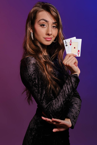 Brünettes Mädchen in schwarzem Samtkleid, das zwei Spielkarten zeigt und seitlich auf buntem Hintergrund posiert. Glücksspiel, Poker, Casino. Nahaufnahme. - Foto, Bild