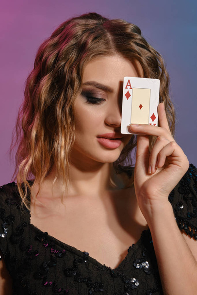 Brown-haired girl in black shiny dress has covered her eye with ace of diamonds, posing on colorful background. Gambling, poker, casino. Close-up. - Foto, Bild