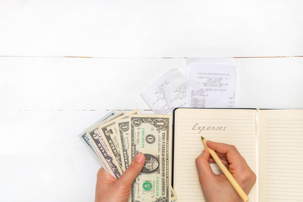 Expenses planning concept. Top view of receipts, woman holding pen and cash and writing words Expenses in a notepad on white wooden table. Image with copy space, flat lay - Photo, Image
