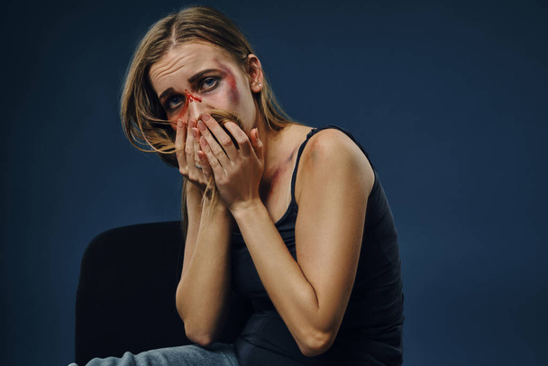 Lijdende vrouw in zwart t-shirt en jeans. Bloedend gezicht met blauwe plekken, bedekt ze het met haar en handpalmen, zittend op een blauwe achtergrond. Close-up - Foto, afbeelding