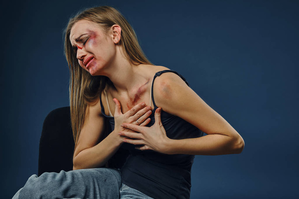 Grieved meisje in zwart t-shirt en jeans. Bloedend gezicht bedekt met blauwe plekken, legde haar handen op de borst en huilde, zittend op een blauwe achtergrond. Close-up - Foto, afbeelding
