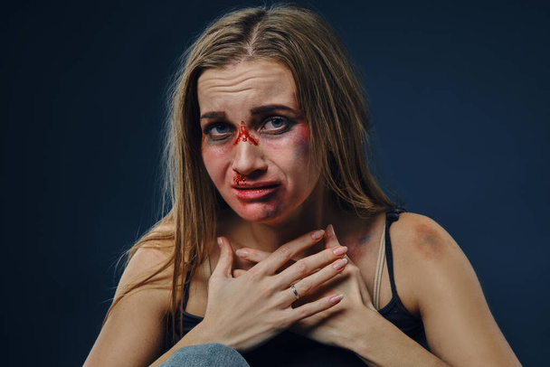 Verängstigte Frau in schwarzem T-Shirt und Jeans. Blutendes Gesicht mit blauen Flecken bedeckt, die Hände auf die Brust gelegt, auf blauem Hintergrund sitzend. Nahaufnahme - Foto, Bild