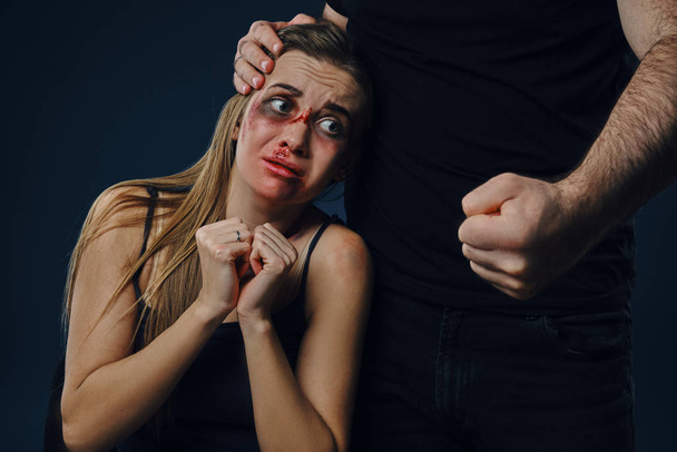 Cruel male put his hand on woman head. Scared victim with bruises on face sitting nearby, terrified. Blue background. Domestic violence. Close-up - Photo, image