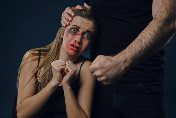 Cruel husband put his hand on wife head. Scared victim with bruises on face sitting nearby, terrified. Blue background. Domestic violence. Close-up - Фото, изображение