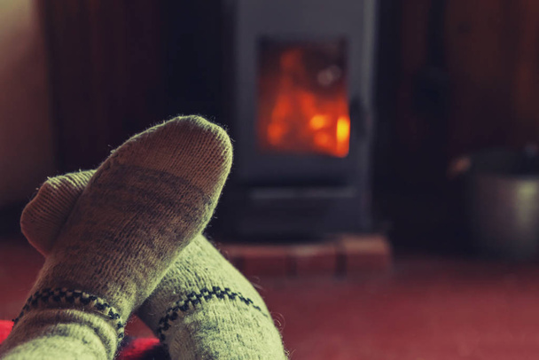 Πόδια σε χειμερινά ρούχα μάλλινες κάλτσες στο φόντο τζάκι. Γυναίκα κάθεται στο σπίτι το χειμώνα ή το φθινόπωρο βράδυ χαλάρωσης και προθέρμανσης. Χειμώνας και κρύο έννοια του καιρού. Παραμονή Χριστουγέννων Hygge. - Φωτογραφία, εικόνα