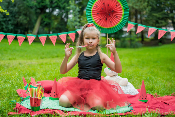 Арбузная вечеринка, пикник для детей в парке. День арбуза. Милая маленькая девочка в шляпе и с игрушечным медведем в арбузной шляпе - Фото, изображение