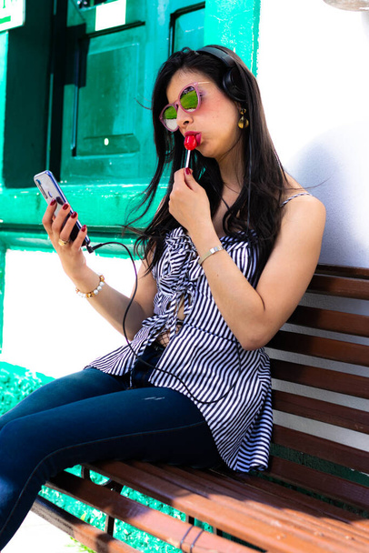 Kolumbiai nő napszemüveget visel, és zenét hallgat a mobiljáról, fejhallgatóval egy fa széken ülve, piros cukorkát eszik. - Fotó, kép