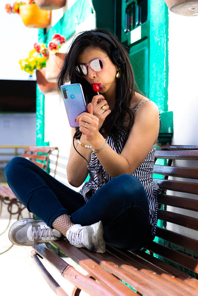 Kolumbijská žena nosí sluneční brýle a poslouchá hudbu z mobilu se sluchátky sedí na dřevěné židli jíst červené bonbóny - Fotografie, Obrázek